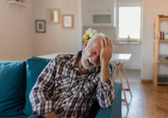 Shot of a senior man with a headache holding his head while sitting on a sofa at home in the living room. Shot of an uncomfortable looking elderly man holding his head in discomfort due to pain at home during the day