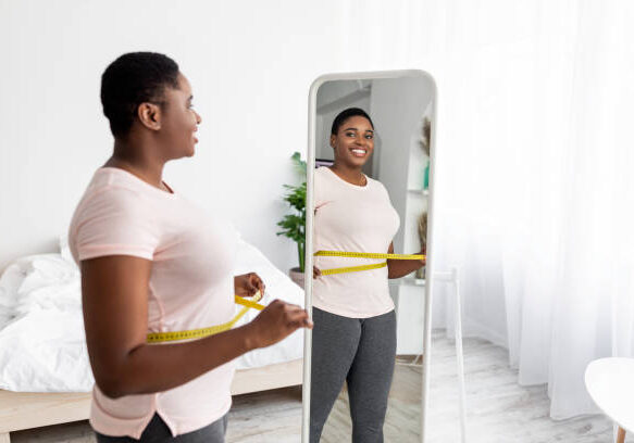 Overweight young black woman measuring her waist with tape near mirror at home, satisfied with slimming result, free space. Plus size African American lady being successful in weight loss