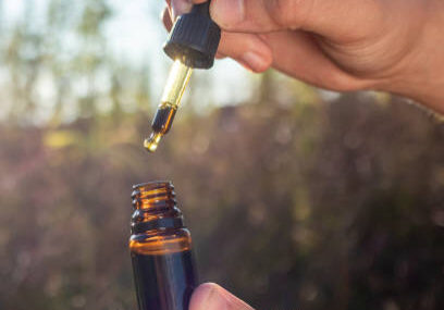 Hands holding and opening CBD oil bottle with dropper pipette, near hemp plant flower bud, in the sunlit field, close up shot.