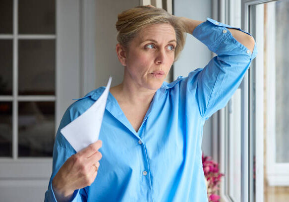 Menopausal Mature Woman Having Hot Flush At Home Cooling Herself With Letters Or Documents