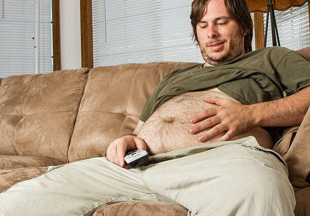 A large belly and a man smiling at it while being lazy with his TV remote in hand. Shows a couch potato or a man with a sedentary lifestyle .