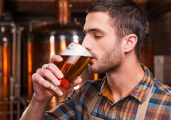 Handsome young male brewer in apron tasting fresh beer and keeping eyes closed while standing in front of metal containers